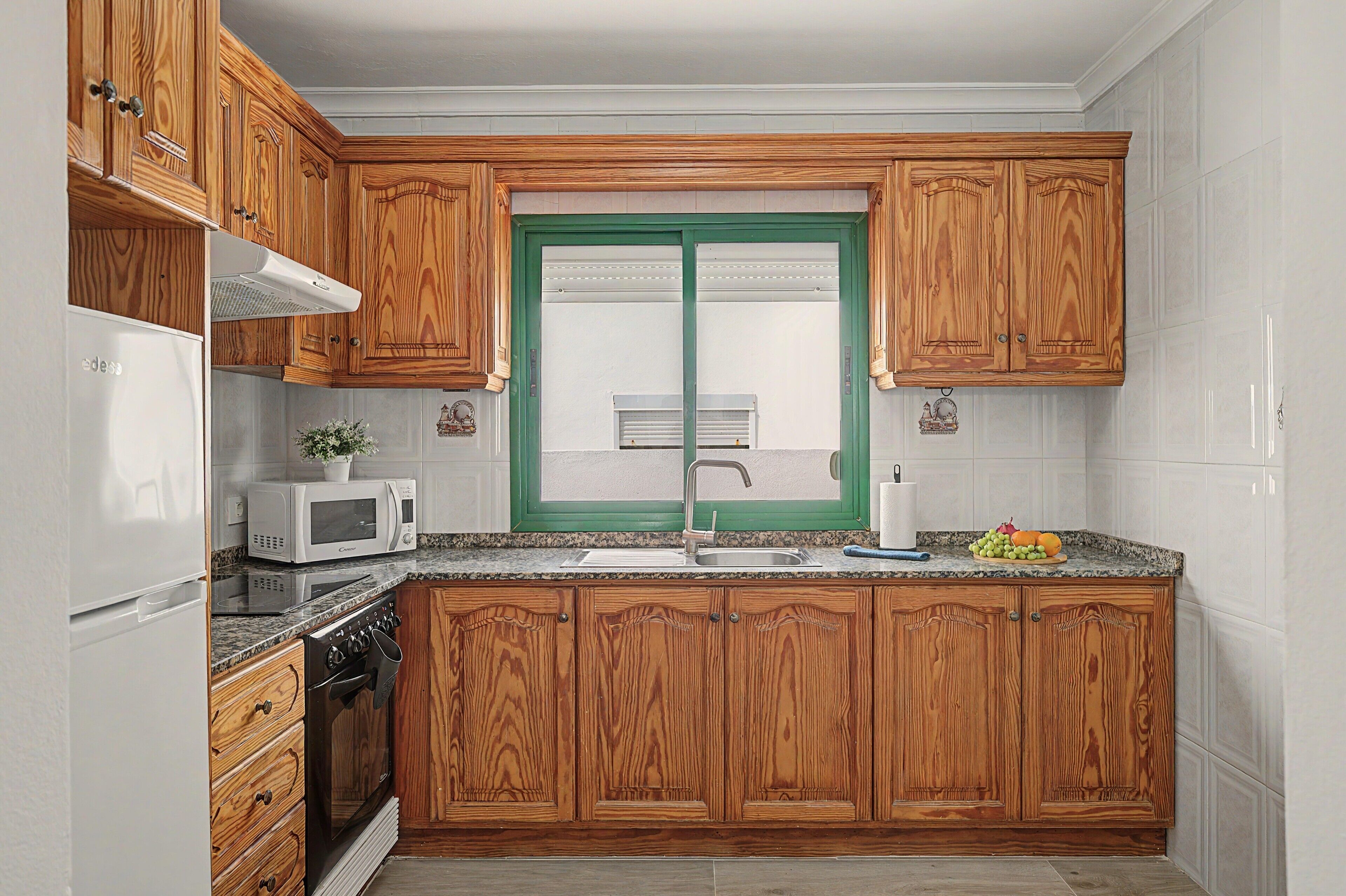 A kitchen with wooden cabinets, a white refrigerator, a microwave, a stove, and a green-framed window.