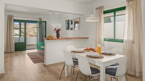 A dining area with a white table set for four, a hanging pendant light, and a view through a window with green shutters.