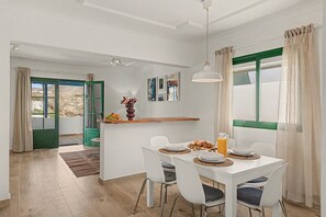 A dining area with a white table set for four, a hanging pendant light, and a view through a window with green shutters.