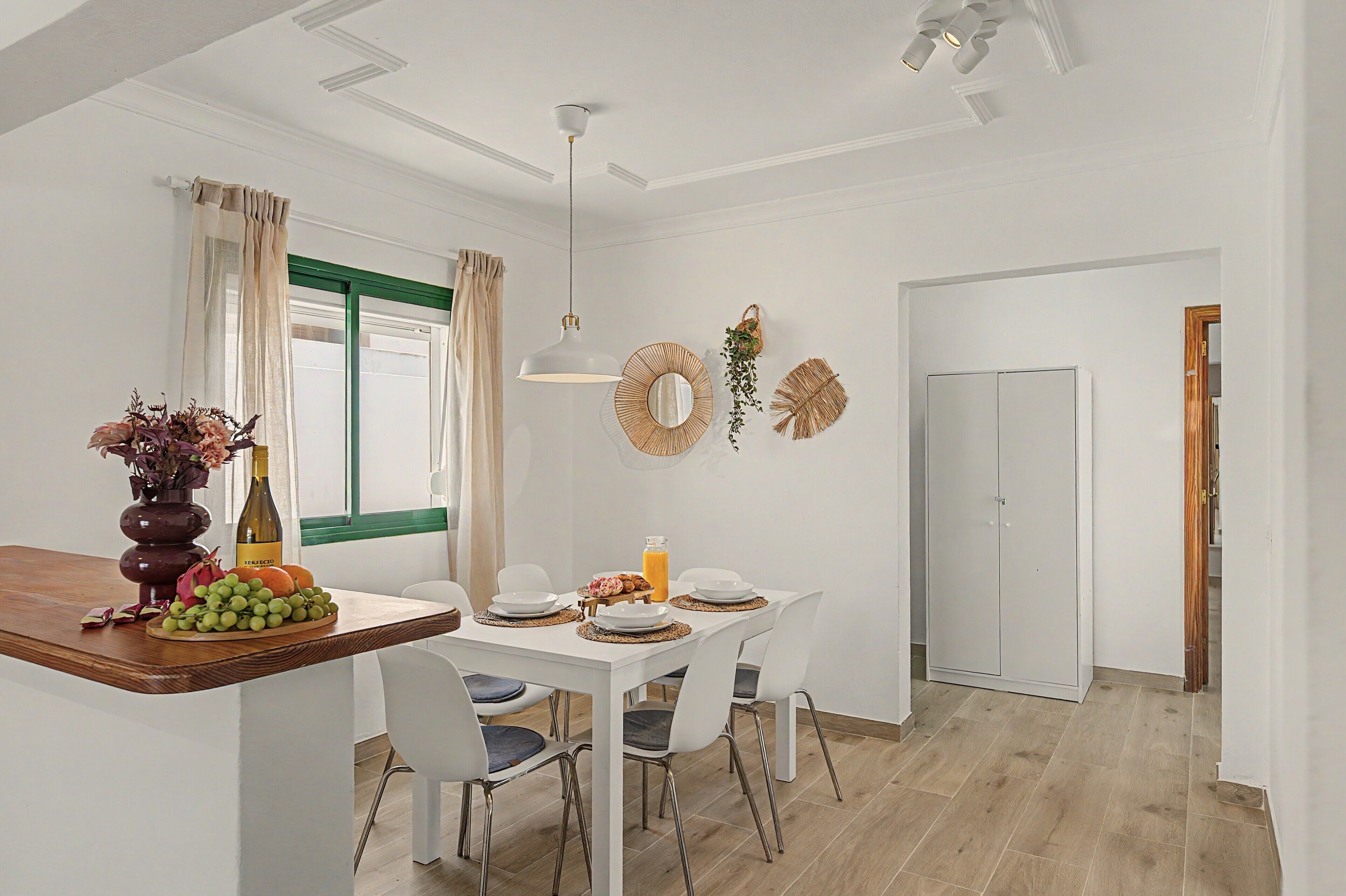 A dining area with a white table set for four, chairs, a vase with flowers, and a bottle of wine.