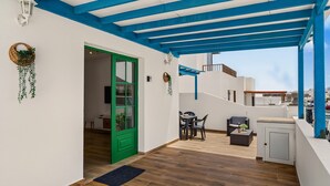 Un espace extérieur abrité, doté d’une table et de chaises, d’une porte verte et d’un mur blanc orné d’une plante suspendue.