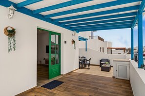 Un espace extérieur abrité, doté d’une table et de chaises, d’une porte verte et d’un mur blanc orné d’une plante suspendue.