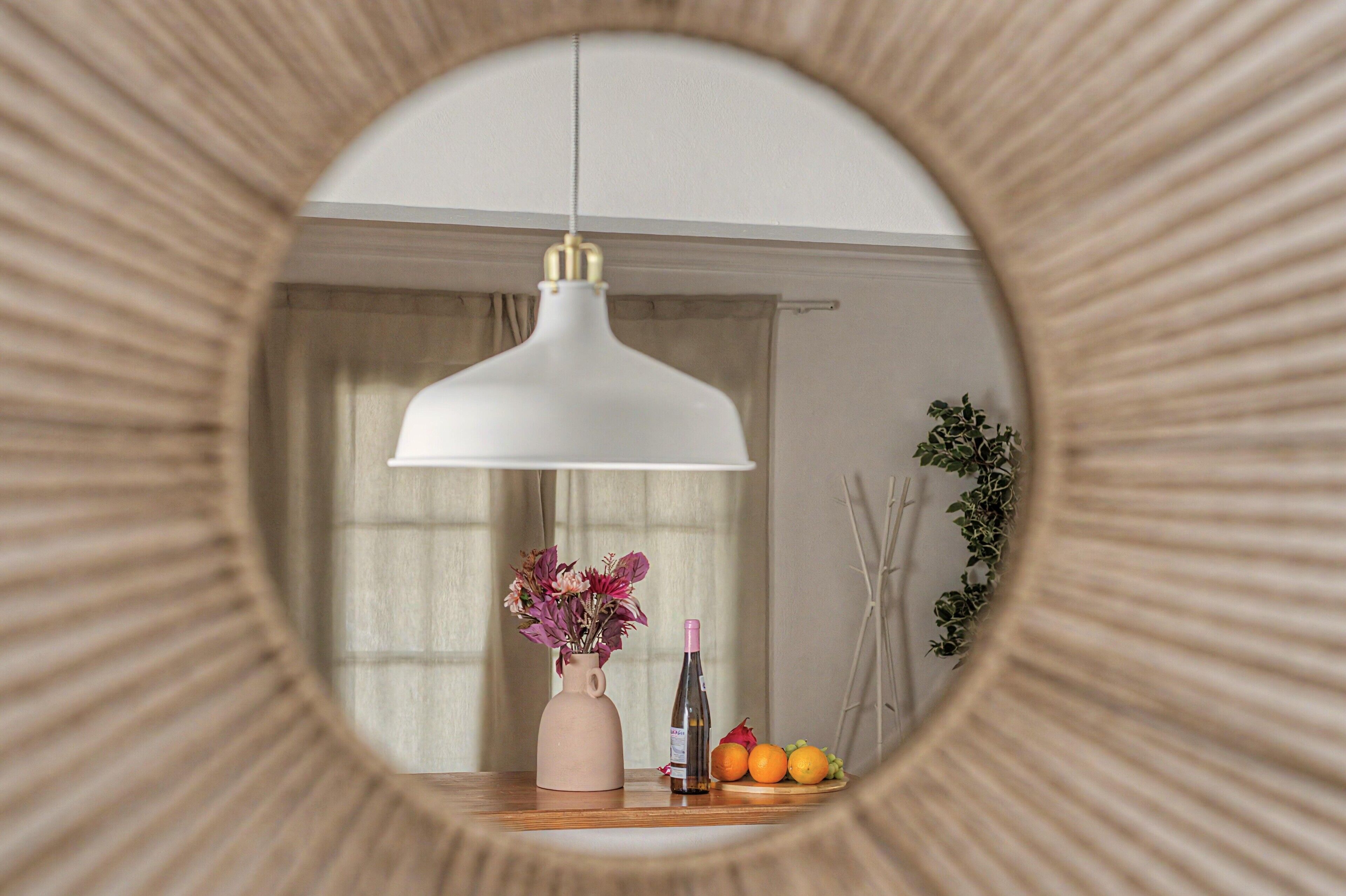A room with a round mirror reflecting a pendant light, a vase with flowers, and a fruit arrangement.