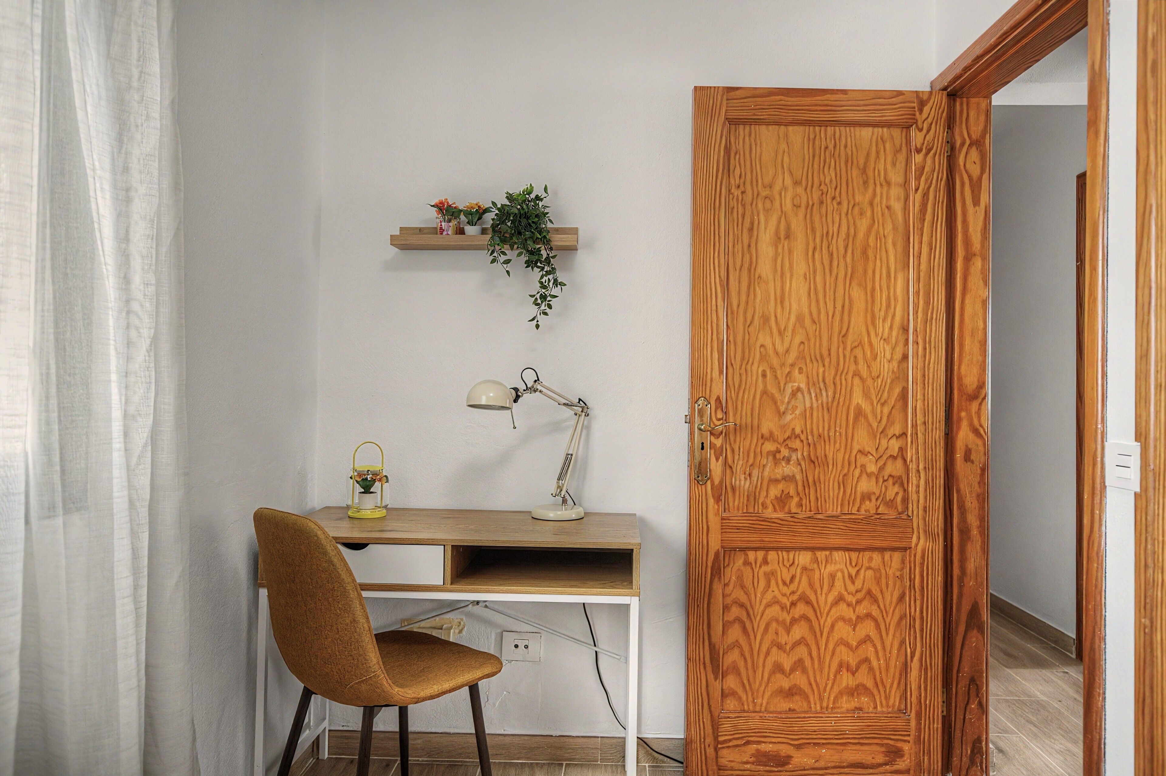 Une pièce avec un bureau en bois, une chaise, une lampe, une étagère avec des fleurs et une porte en bois.