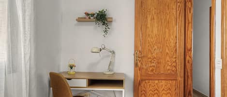 Une pièce avec un bureau en bois, une chaise, une lampe, une étagère avec des fleurs et une porte en bois.