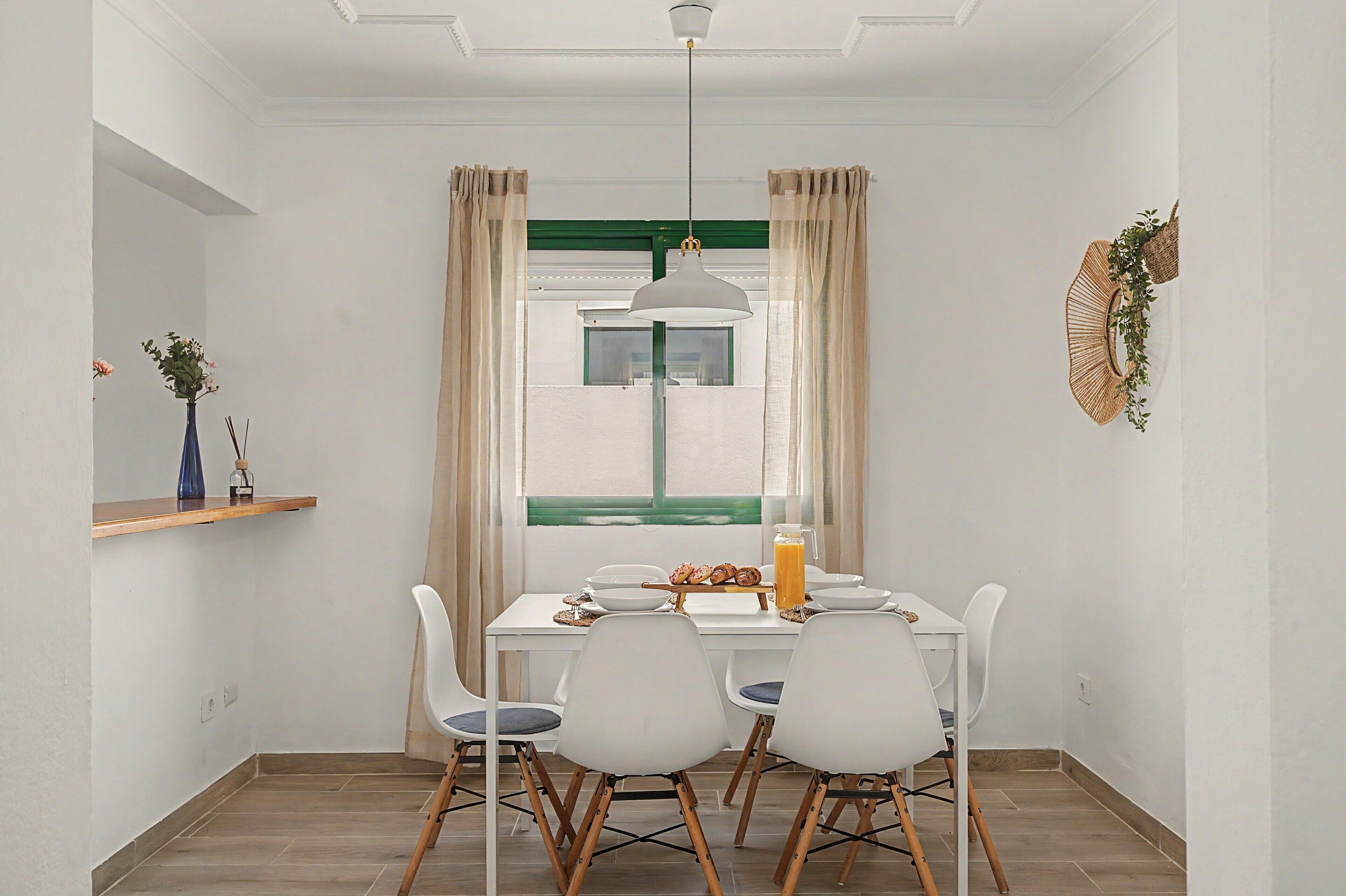 Comedor con mesa blanca, cuatro sillas, lámpara colgante y ventana con cortinas.