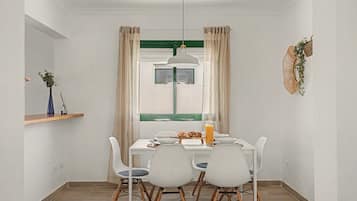 Comedor con mesa blanca, cuatro sillas, lámpara colgante y ventana con cortinas.