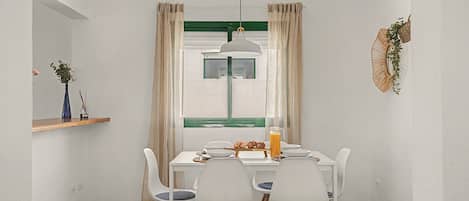 Un coin repas avec une table blanche, quatre chaises, un luminaire suspendu et une fenêtre avec des rideaux.