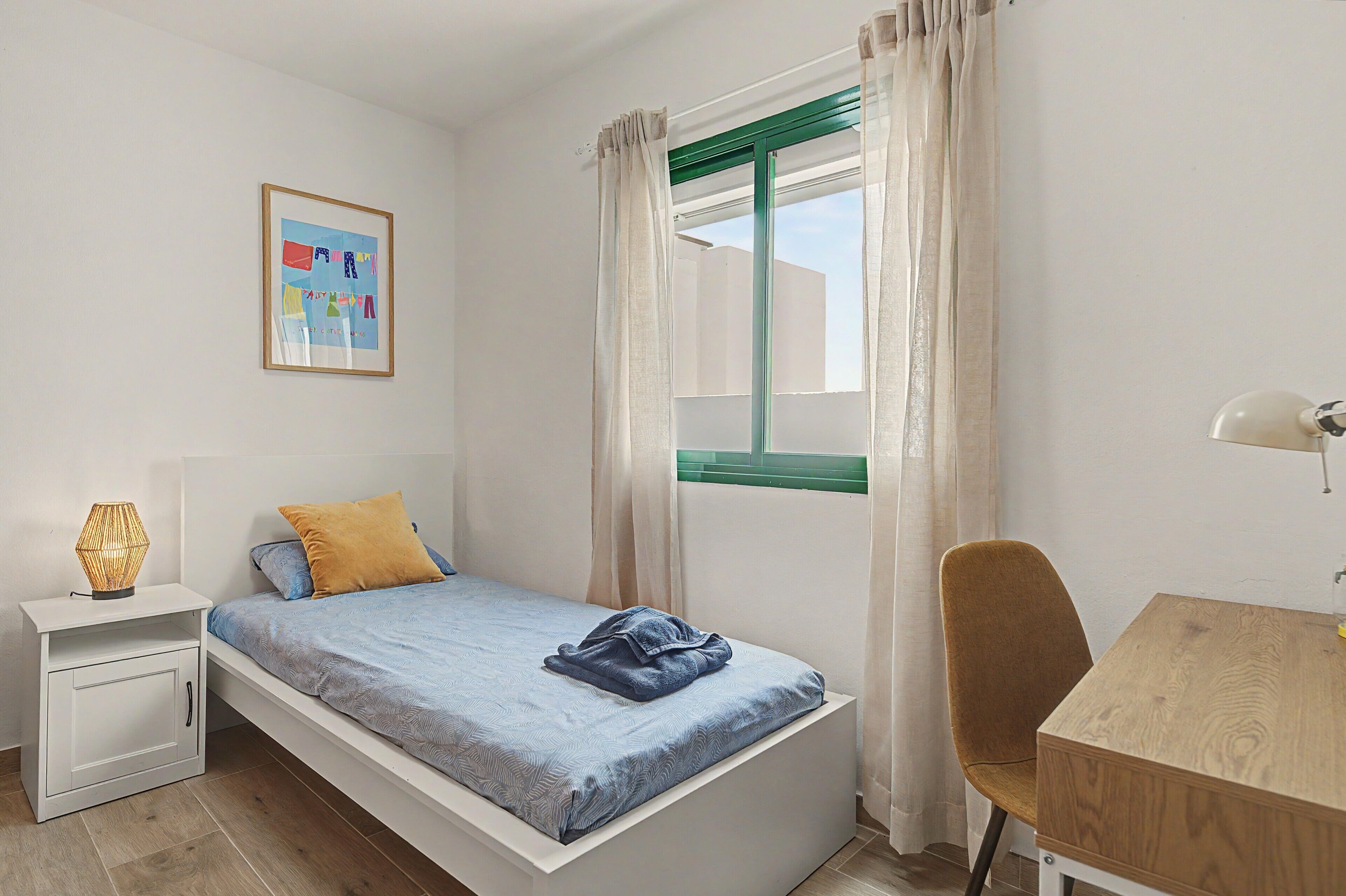 A small, neatly arranged bedroom with a single bed, a desk, a chair, and a bedside lamp.