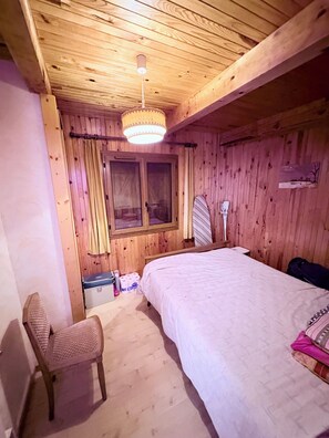 Ein Zimmer mit Holzboden, einem Bett, einem Sessel, einem Fenster mit Vorhängen und einer Hängeleuchte.