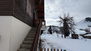 Un edificio de madera con una escalera que lleva a una entrada con rejas, rodeado de un paisaje nevado y árboles.