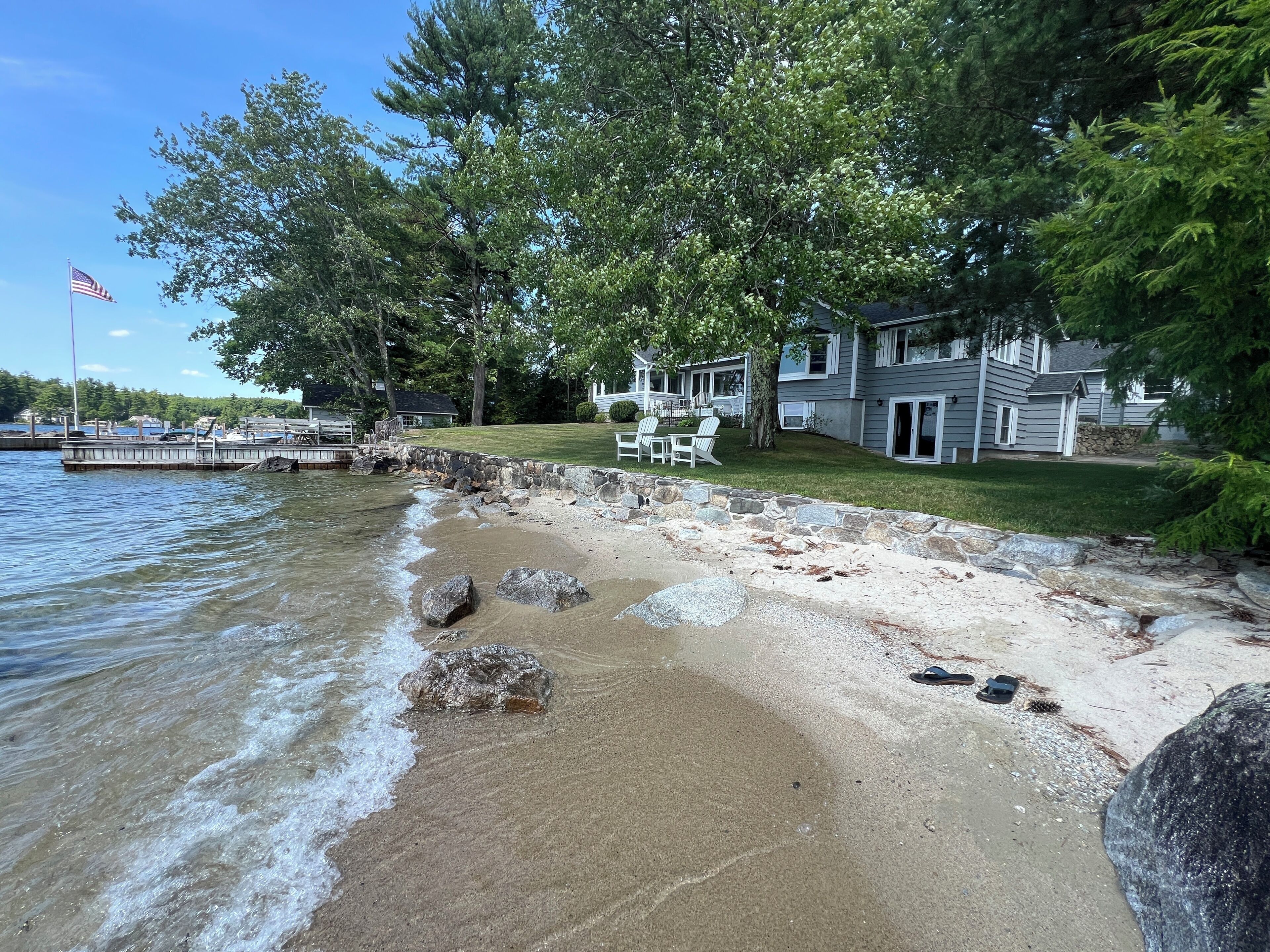 A lakeside property with a stone beach, a dock, and a house with a lawn and outdoor seating.