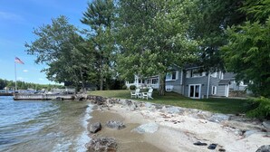 A lakeside property with a stone beach, a dock, and a house with a lawn and outdoor seating. - Exceptional Lakefront Home on Springfield Point – Lake Winnipesaukee (Wolfeboro)