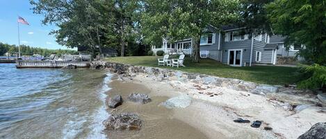 A lakeside property with a stone beach, a dock, and a house with a lawn and outdoor seating.