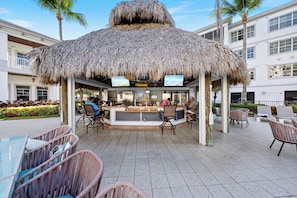 A thatched-roof outdoor bar area with seating and a counter, surrounded by palm trees and a building with multiple windows. - Beautiful, Stylish Poolside Condo! (Bonita Springs)