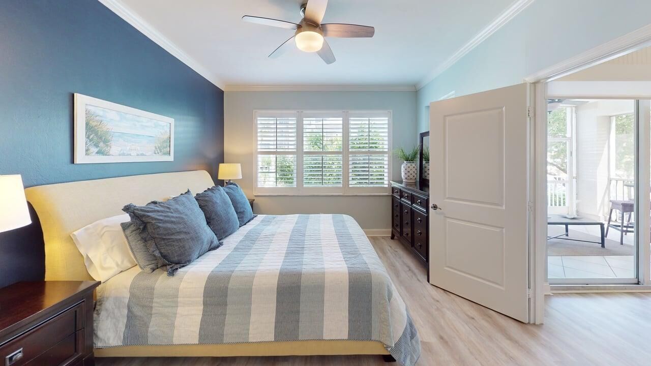 A bedroom with a bed, a ceiling fan, a window with blinds, a door leading to a balcony, and a nightstand with a lamp.
