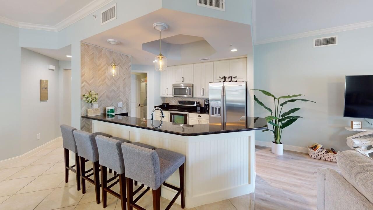 A modern kitchen with white cabinets, a central island with bar stools, a microwave, a refrigerator, and a potted plant.