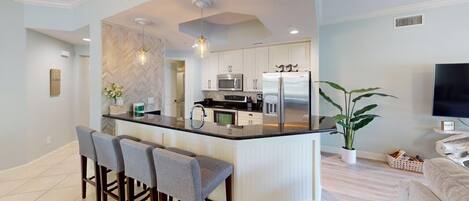 A modern kitchen with white cabinets, a central island with bar stools, a microwave, a refrigerator, and a potted plant.