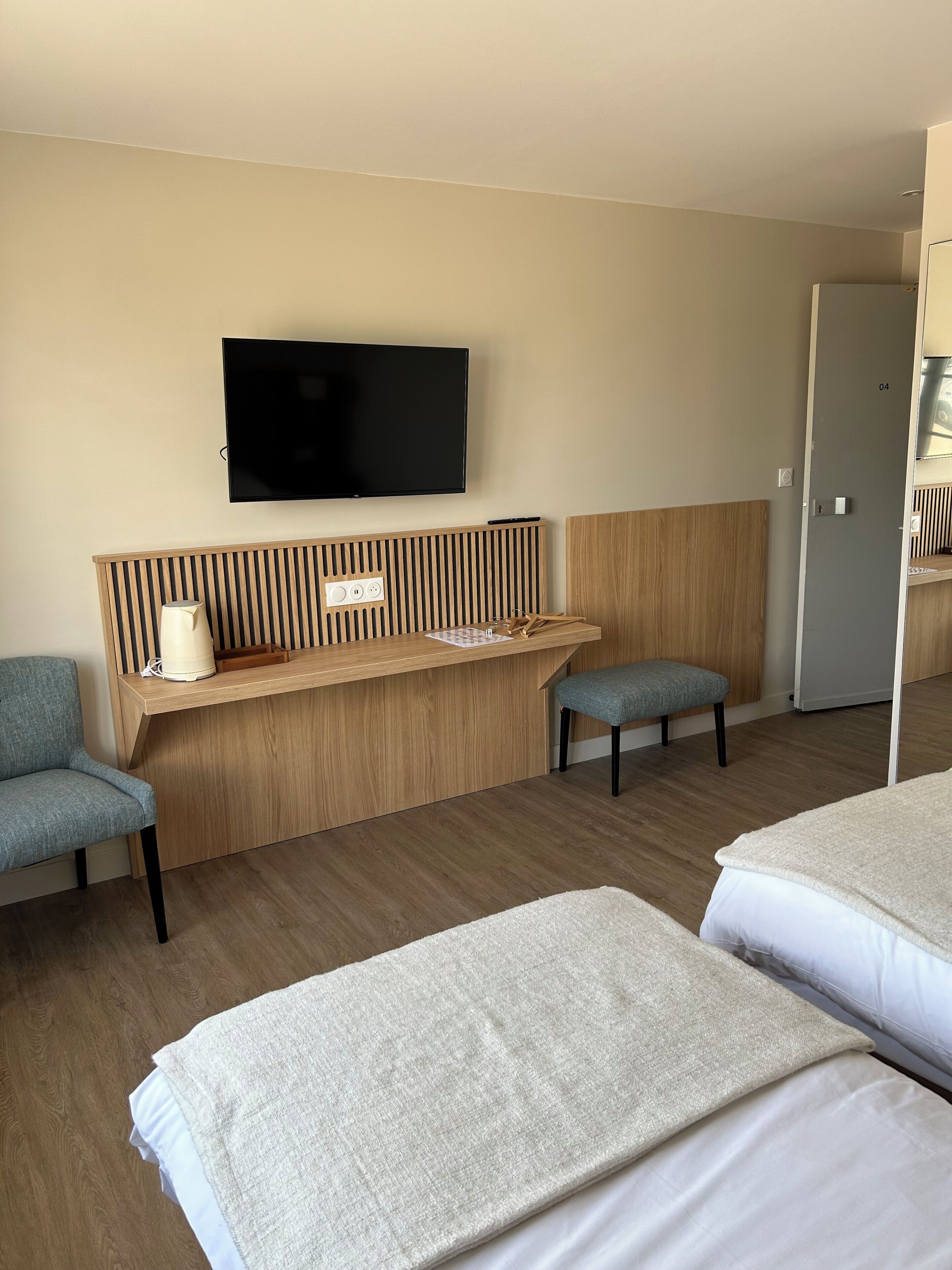 Habitación de hotel con televisión en la pared, cabecera de madera, escritorio con l ámpara, dos sillas y dos camas.