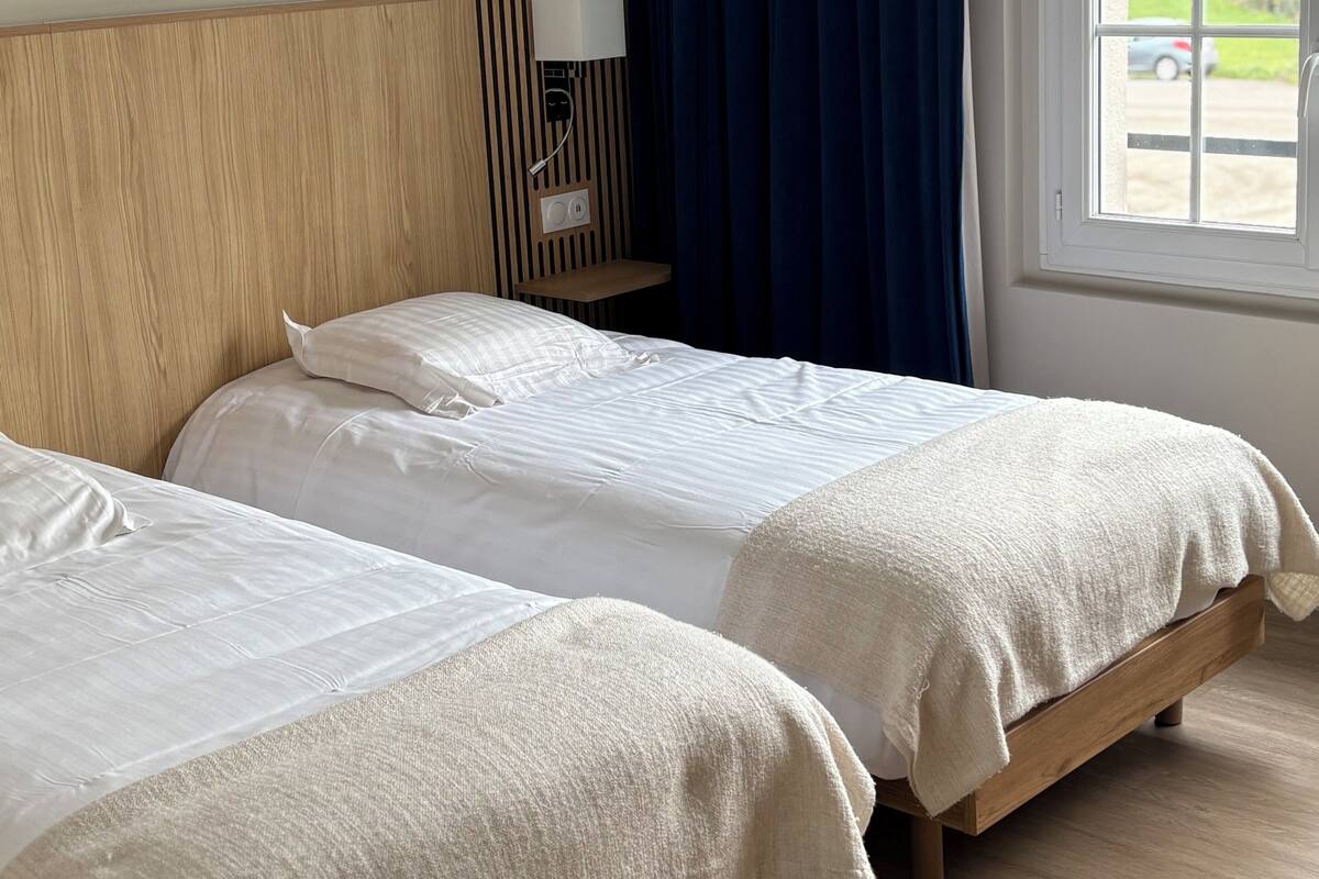 Ein Hotelzimmer mit zwei Betten, einem Holzkopfstück, einem Fenster mit Vorhängen und Blick ins Freie.