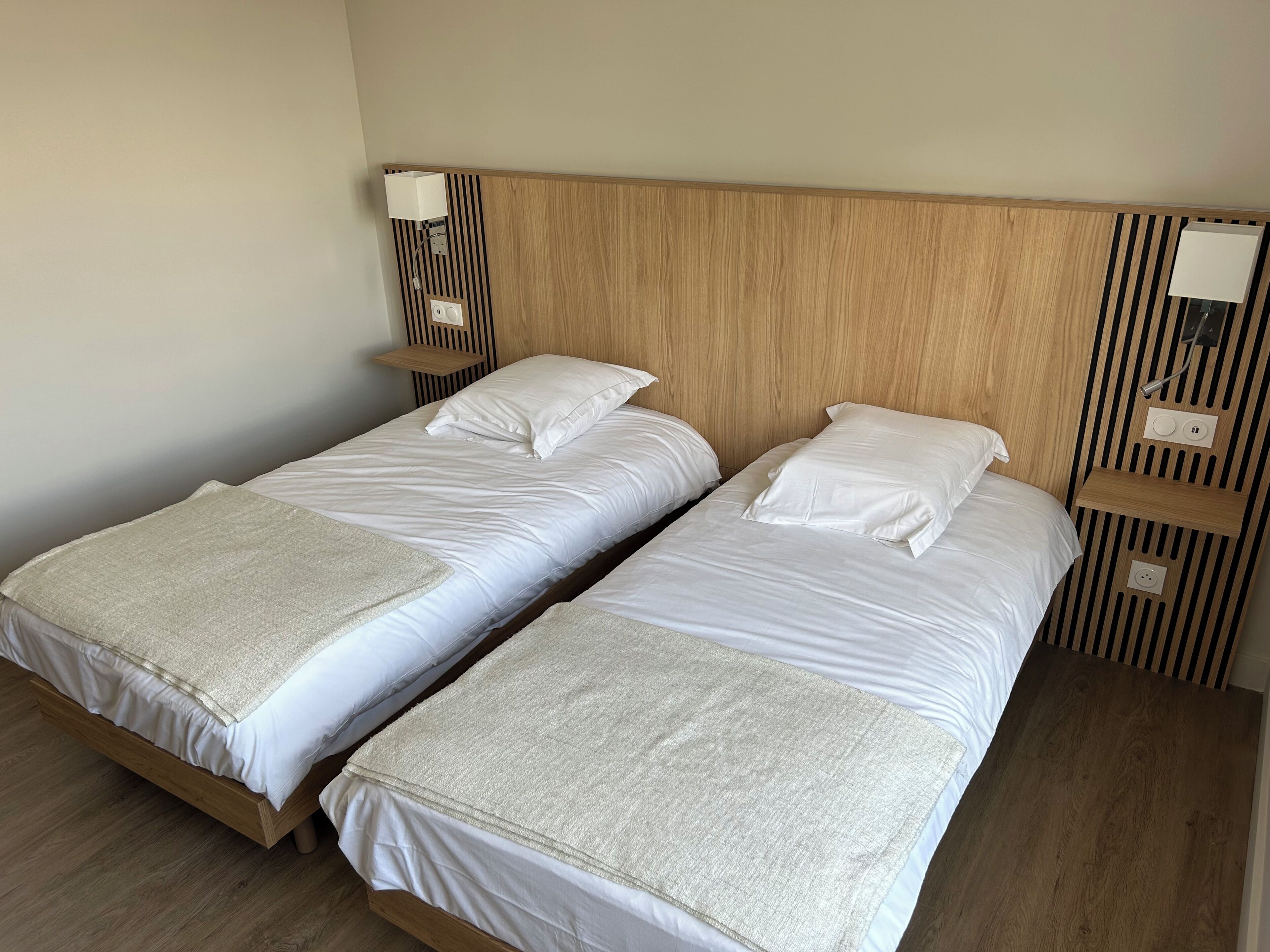 Two single beds with white linens and beige throw blankets, wooden headboards, and a wall-mounted lamp.