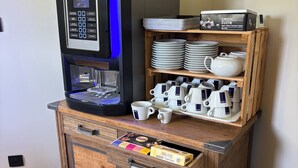 A coffee machine on a wooden cabinet with a shelf above it holding plates, cups, and a teapot. - Hôtel L'Escale des Hayons (Esclavelles)