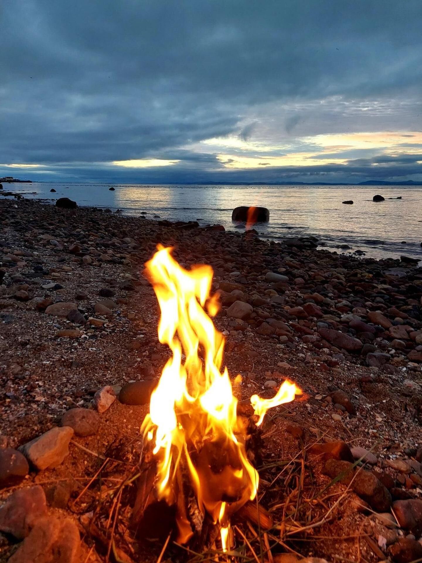 A campfire on a rocky beach with a view of the ocean and distant land.