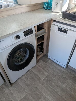 A compact kitchen with a washing machine, dishwasher, and wooden shelves. - Seaside Escape Lodge in Port Seton Sands (Prestonpans)