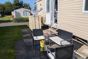 A mobile home with a patio area featuring two chairs and a small table, a yellow bucket, and a clothesline with laundry. - 2 Bed - Sleeps 4 - Seton Sands Park (Prestonpans)