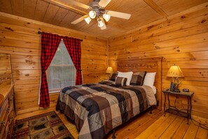 A wooden cabin bedroom with a bed, two bedside lamps, a ceiling fan, and a window with red and black checkered curtains. - Hot Tub | Fireplace | Multicade | Mountain Views (Sevierville)
