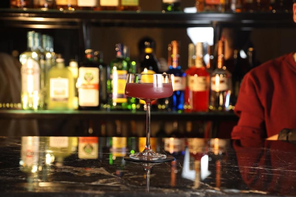 A bar setting with a glass of red cocktail on a reflective surface, surrounded by various bottles of liquor.