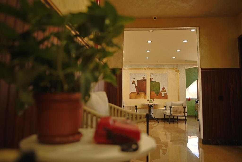 Un vestíbulo de hotel con una planta en maceta, una maleta roja y una vista parcial de un comedor con mesa y sillas.