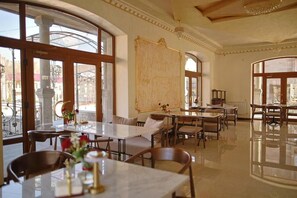 A spacious dining area with multiple tables and chairs, large windows offering views of the street, and a decorative wall panel. - Passage Hotel Restaurant (Goris)