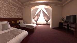 A hotel room with two beds, a TV, a desk, and a large window with curtains. - Passage Hotel Restaurant (Goris)