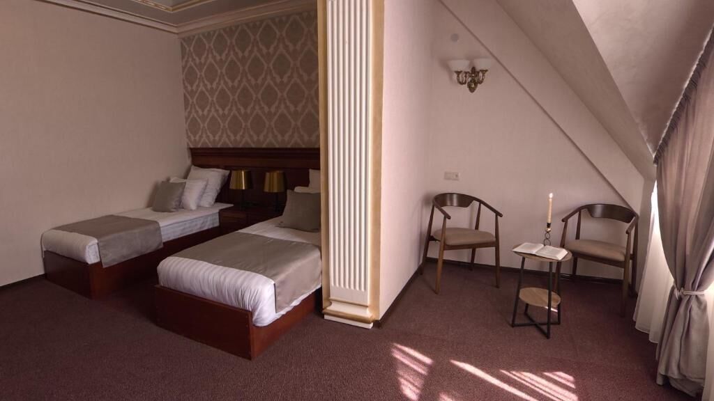 Ett hotellrum med två separata sängar, en sittgrupp med en stol och ett bord samt ett stort fönster som släpper in naturligt ljus.