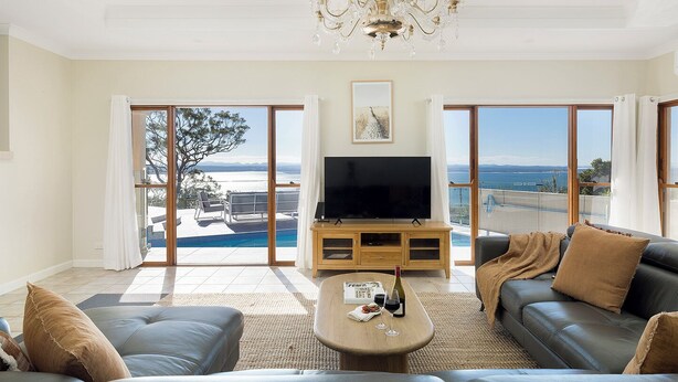 Ein Wohnzimmer mit Fernseher, Sofa und Couchtisch, große Schiebeglasfenster zu einem Balkon mit Blick auf einen Pool.