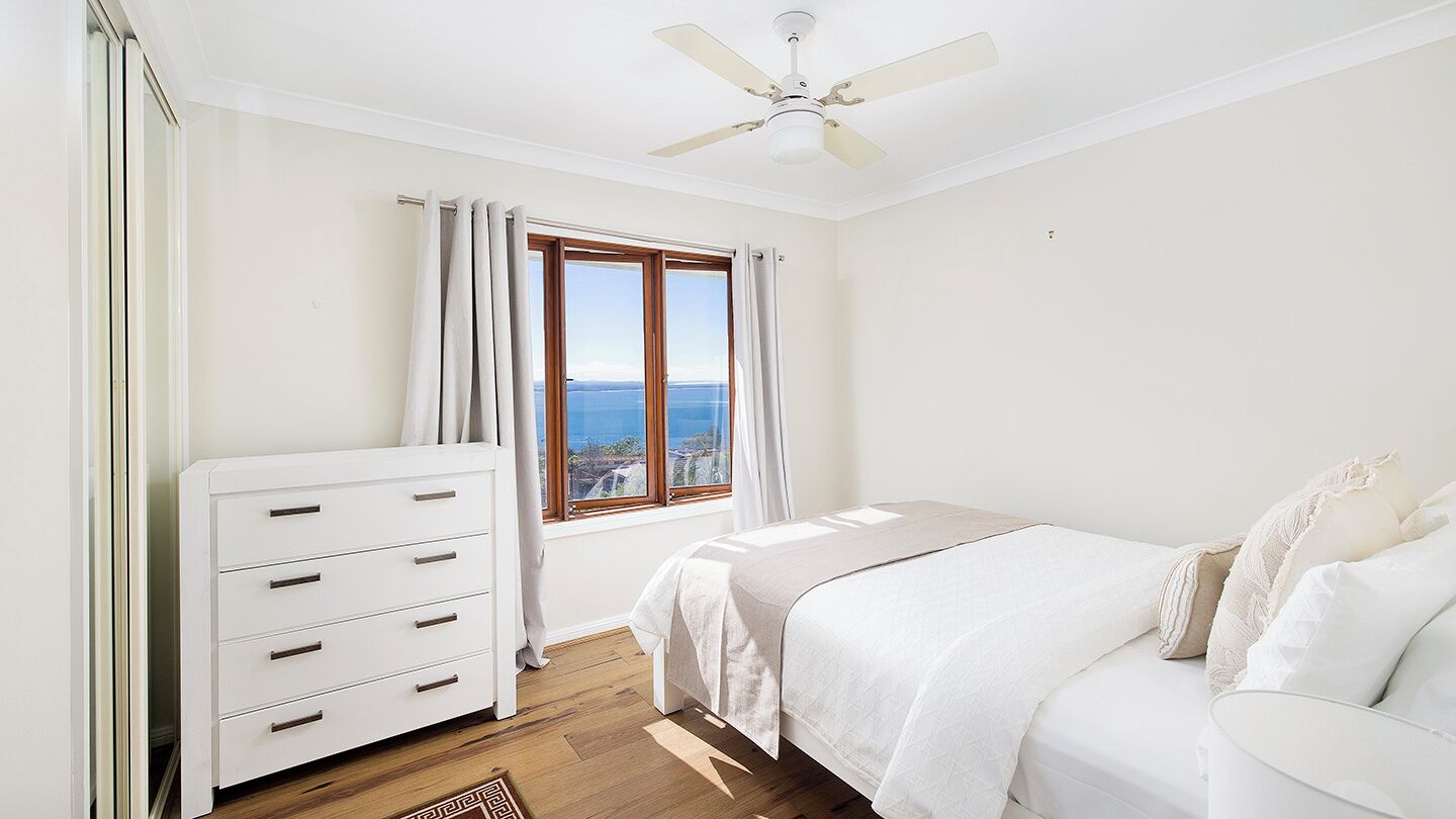 A bedroom with a bed, a white dresser, a ceiling fan, a window with curtains, and a view of the sea.