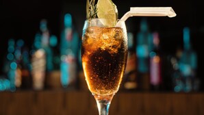 Et glas med en mørk drik med is, pyntet med et skives citron og en rosmarin kvist, samt et sugerør, stående på en træbar.