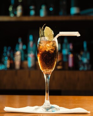 A glass of a dark-colored beverage with ice, garnished with a slice of lime and a sprig of rosemary, with a straw, on a wooden bar counter. - Kasian Hotel (Addis Ababa)