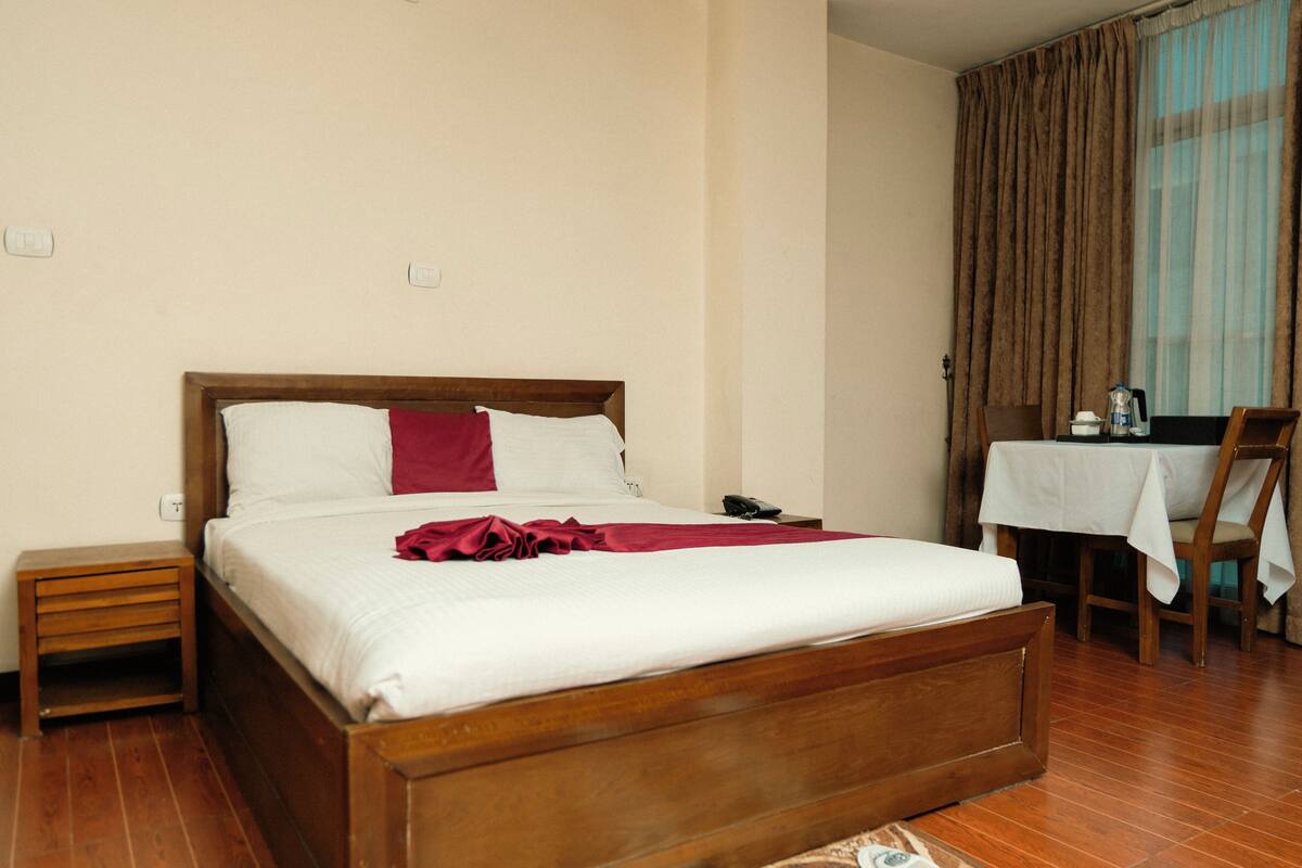 Habitación de hotel con una cama de madera, una mesita de noche pequeña, una silla y una mesa con mantel blanco.