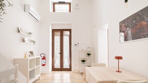 A bright, white room with a tiled floor, a white table with a red lamp, a white shelf with plants, a white cabinet, and a door with frosted glass panels. - Apartment 'L'angolo Del Teatro' with Private Terrace and Air Conditioning (Galatone)