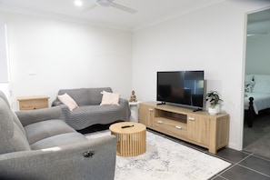 Una sala de estar moderna con un sofá gris, un mueble de televisión de madera, un control remoto sobre una mesa redonda de madera y una pequeña estatua.