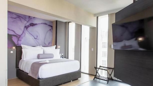 Une chambre moderne avec un grand lit, un mur d’accent violet et une vue sur un bâtiment à l’extérieur.