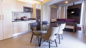 Une cuisine moderne avec un coin repas comprenant une table et des chaises en verre, ainsi qu’un salon avec un canapé et un téléviseur.