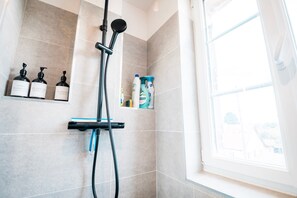 Une douche équipée d’un pommeau de douche à main, d’un flexible de douche et de bouteilles de produits de toilette disposées sur une étagère.