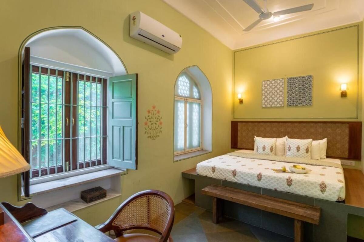 Una habitación con cama, escritorio, silla, ventana con rejas, aire acondicionado y ventilador de techo.