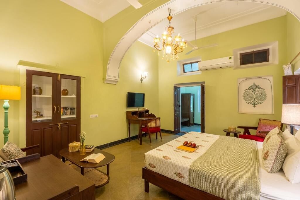 A bedroom with a bed, a desk, a chair, a TV, and a door leading to another room.