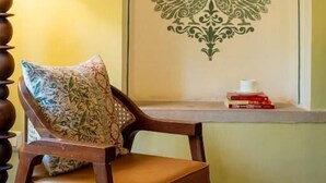 Een kamer met een houten stoel, een gestreept kussen, een tafel met een decoratief object aan de muur, en een lamp.