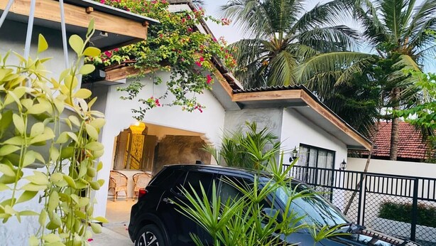 A black SUV parked in a driveway, a white building with a wooden roof, and a fenced area with palm trees.