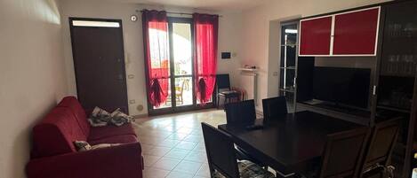 A living room with a red sofa, a dining table with chairs, a TV, and a kitchen area with cabinets.